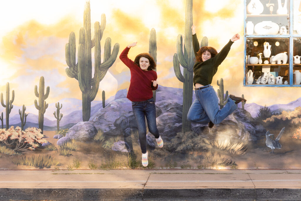 Highschool sisters jumping in front of a cactus mural during a sister session in Downtown Tucson. Katie Gilbert Photography.