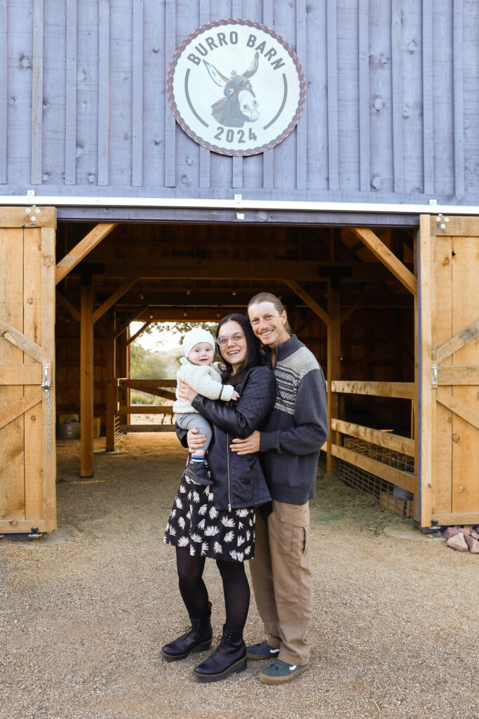 A family of 3 standing in front of a blue rustic barn. 