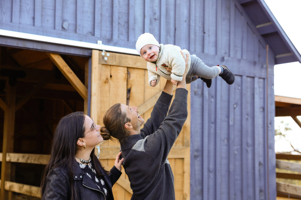 Family holding their baby in the air in front of a rustic barn.